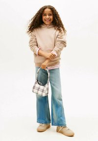 Ragazza sorridente con capelli ricci che indossa una felpa beige con cappuccio, jeans a gamba larga blu, sneakers beige, e tiene in mano una piccola borsa a quadri, in piedi su uno sfondo semplice.