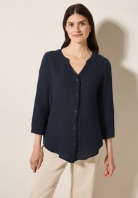 Camicia blu navy con bottoni, dotata di un motivo testurizzato, maniche a tre quarti e orlo curvo. Il colletto è arrotondato e presenta una staffa di bottoni.