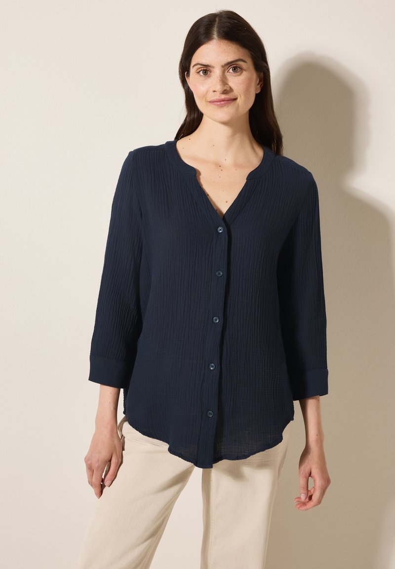 Camicia blu navy con bottoni, dotata di un motivo testurizzato, maniche a tre quarti e orlo curvo. Il colletto è arrotondato e presenta una staffa di bottoni.