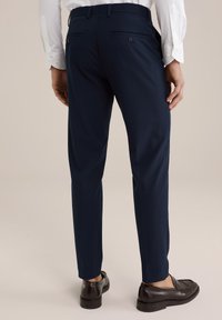 Pantalon de costume marine avec une coupe ajustée, doté de deux poches arrière et d'une texture de tissu lisse. Porté avec des mocassins noirs.