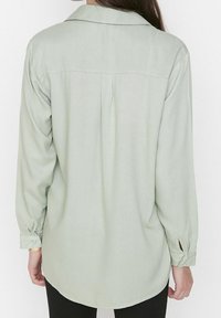 Light green button-up shirt with long sleeves, rounded hem, back yoke detail, and tied cuffs. Smooth texture and casual fit.
