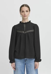 Blusa nera a maniche lunghe con scollatura e polsini arricciati. Presenta un giromanica frontale con pannello testurizzato e dettagli in pizzo. Tessuto liscio, vestibilità comoda.