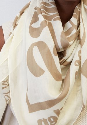 Écharpe crème clair avec de grands motifs abstraits beige, drapée autour du cou et des épaules, portée sur un haut tricoté crème.