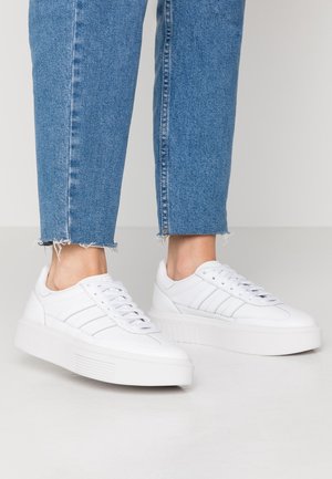 Białe trampki z gładką skórzaną cholewką, teksturowanymi bocznymi paskami i grubą białą gumową podeszwą. Połączone z wyblakłymi niebieskimi dżinsami z postrzępionymi nogawkami.