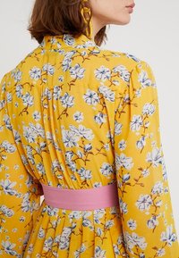 Robe florale jaune à manches longues, dotée d'une ceinture en tissu rose. Le design comprend des fleurs bleues et blanches avec un tissu doux et texturé.