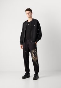 Black bomber jacket with zip, plain black T-shirt, patterned black sweatpants with a graphic, and black sneakers. Simple, relaxed fit.