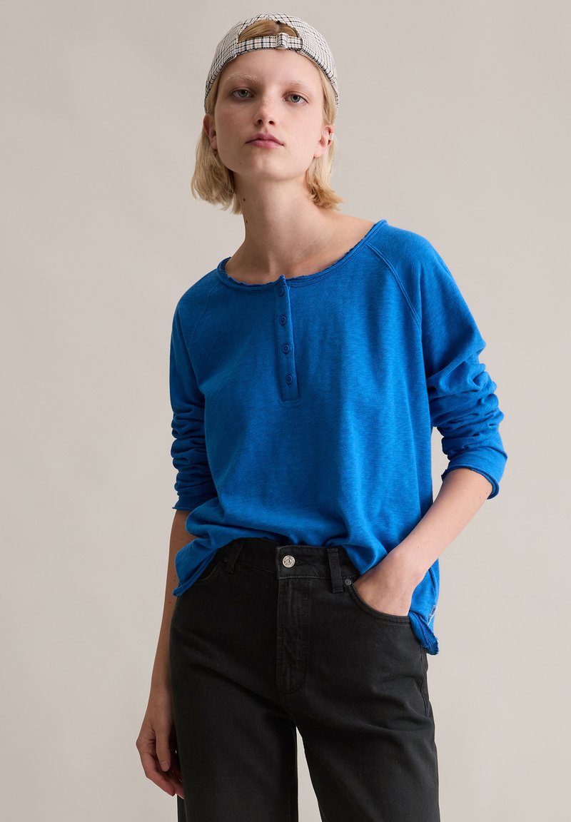 Niebieska koszula henley z długim rękawem, okrągłym dekoltem i trzema guzikami, w połączeniu z czarnymi dżinsami. Gładka tekstura i luźny krój.