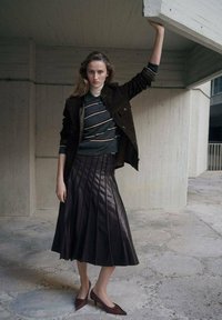 Falda plisada de cuero marrón, top a rayas verdes oscuras y chaqueta marrón, complementados con tacones burdeos. Texturas suaves y diseño estructurado.