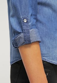 Camicia in denim con maniche arrotolate e chiusura a scatto, di colore azzurro chiaro e leggere variazioni di texture. I bordi sono leggermente sfilacciati.