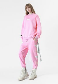 Sudadera rosa y pantalones de chándal a juego, hechos de una tela suave. Presenta una etiqueta en el pecho y detalles decorativos. Combinados con botas blancas.