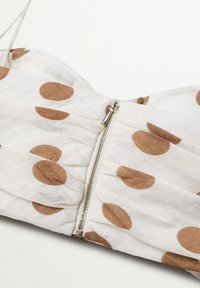 Tissu blanc à pois marron avec une fermeture éclair en métal argenté au centre, avec des coutures froncées de chaque côté de la fermeture.