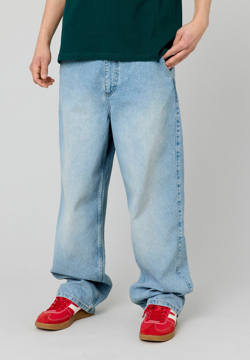 Jean en denim bleu clair, baggy, avec un délavage léger, arborant un design traditionnel à cinq poches. Associé à des baskets rouges.