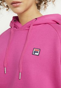 Růžová mikina s texturovanou tkaninou, stažitelnou kapucí a malou vyšitou nášivkou v červené, bílé a modré barvě na hrudi.