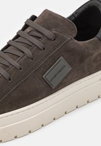 Hnědé semišové tenisky s tmavohnědými tkaničkami, černým vnitřním podšitím a bílou texturovanou podrážkou, které mají na boku obdélníkovou logo nálepku.