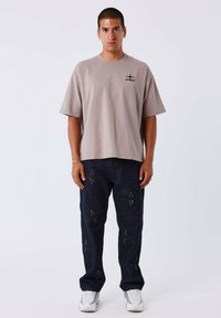 Camiseta de algodón beige oversized con un logo impreso, combinada con pantalones vaqueros oscuros con diseños de costura en forma de estrella y zapatillas deportivas blancas.