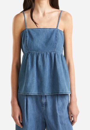 Kvinde iført en ærmeløs blå denimtop med tynde stropper og løstsiddende blå denimbukser mod en ensfarvet baggrund.