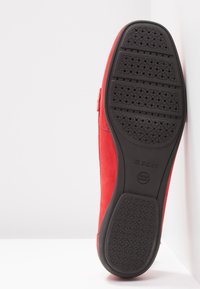 Chaussure en suède rouge avec une semelle plate en caoutchouc noir dotée d'un motif texturé. Le côté présente une sangle en Velcro pour sécuriser la chaussure.
