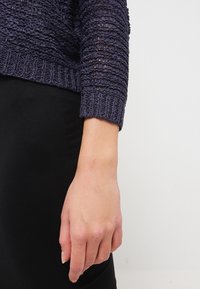 Suéter de punto azul marino con patrón texturizado y puños acanalados. Mostrado de lado, combinado con una prenda negra.