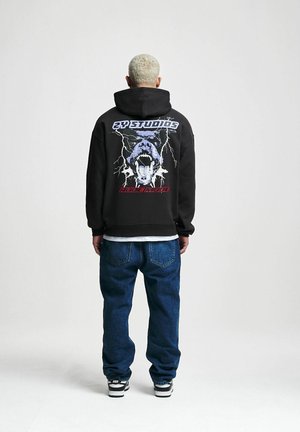 Black hoodie with graphic design featuring a snarling face and lightning bolts. Paired with blue jeans and black and white sneakers.