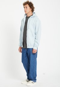 Sweat à zip bleu clair avec deux poches avant, superposé sur un t-shirt gris foncé. Porté avec un jean bleu ample et des baskets blanches.