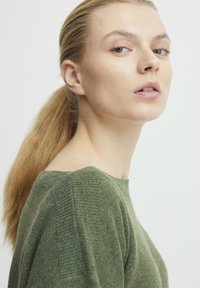 Grüner Strickpullover mit weitem Ausschnitt und gerippter Textur. Das Model hat langes, zurückgebundenes Haar, vor einem neutralen Hintergrund.