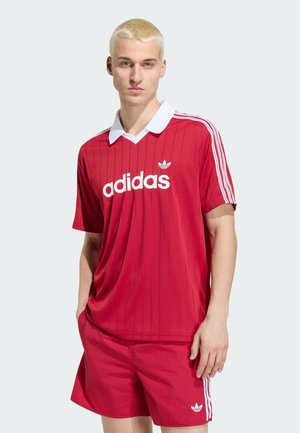 Męski model z krótkimi blond włosami, ubrany w czerwoną sportową koszulkę Adidas i pasujące spodenki z białymi paskami i logo na jednolitym tle.