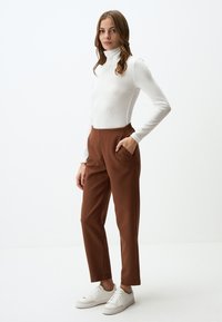 Maglione a collo alto bianco a coste, pantaloni marroni a sigaretta con tasche laterali e sneakers bianche su uno sfondo neutro. Texture lisce e design aderente.