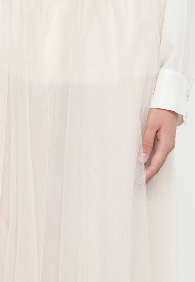 Lichtgewicht, gelaagde tule jurk in zacht ivoor. Heeft een delicate textuur, subtiele doorschijnendheid en een aangesloten taille met een knoopdetail aan de manche.