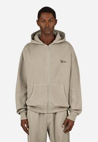 Beige glidlås hettegenser laget av ribbestrikket stoff, med en frontlomme, hette og subtil brodering på brystet. Myk tekstur og avslappet passform.