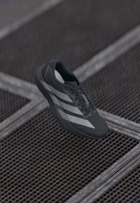 Šedá sportovní bota s texturovaným svrškem, zdobená třemi bílými pruhy, tkaničkami a lehkým designem, vystavená na pozadí z kovové mřížky.
