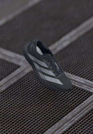 Grauer Sportschuh mit strukturiertem Obermaterial, ausgestattet mit drei weißen Streifen, Schnürsenkeln und einem leichten Design, vor einem metallenen Gitterhintergrund präsentiert.