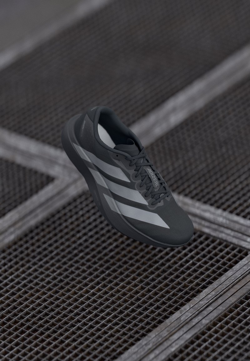 Šedá sportovní bota s texturovaným svrškem, zdobená třemi bílými pruhy, tkaničkami a lehkým designem, vystavená na pozadí z kovové mřížky.