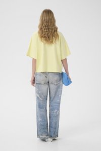 Kvinde med bølget blondt hår iført en løs gul skjorte, falmede vide jeans, hvide hæle, holder en blå, fluffy clutch, set bagfra.