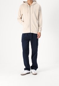 Beige zip-up hoodie med dragsko, framfickor; kombinerad med marinblå byxor och vita sneakers med grå sulor. Enkla, avslappnade design.