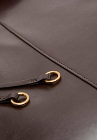 Superficie in pelle marrone con una texture liscia, con anelli in metallo dorato e cinturini in pelle scura, accentuata da un'elegante cucitura.