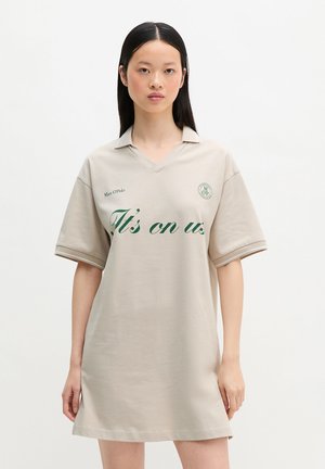 Joven mujer que lleva un vestido polo beige de gran tamaño con texto verde "It's on us" y el logo de la marca, de pie sobre fondo blanco.