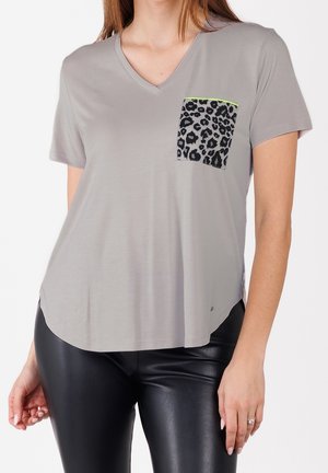 Kobieta w jasnoszarej koszulce z dekoltem w serek, krótkim rękawem i przednią kieszenią w panterkę, w połączeniu z czarnymi spodniami z ekologicznej skóry.