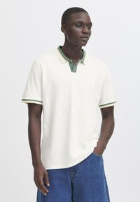 Camisa polo blanca con detalles en verde y amarillo, con cuello abotonado y mangas cortas. Llevada con pantalones cortos de mezclilla azules.