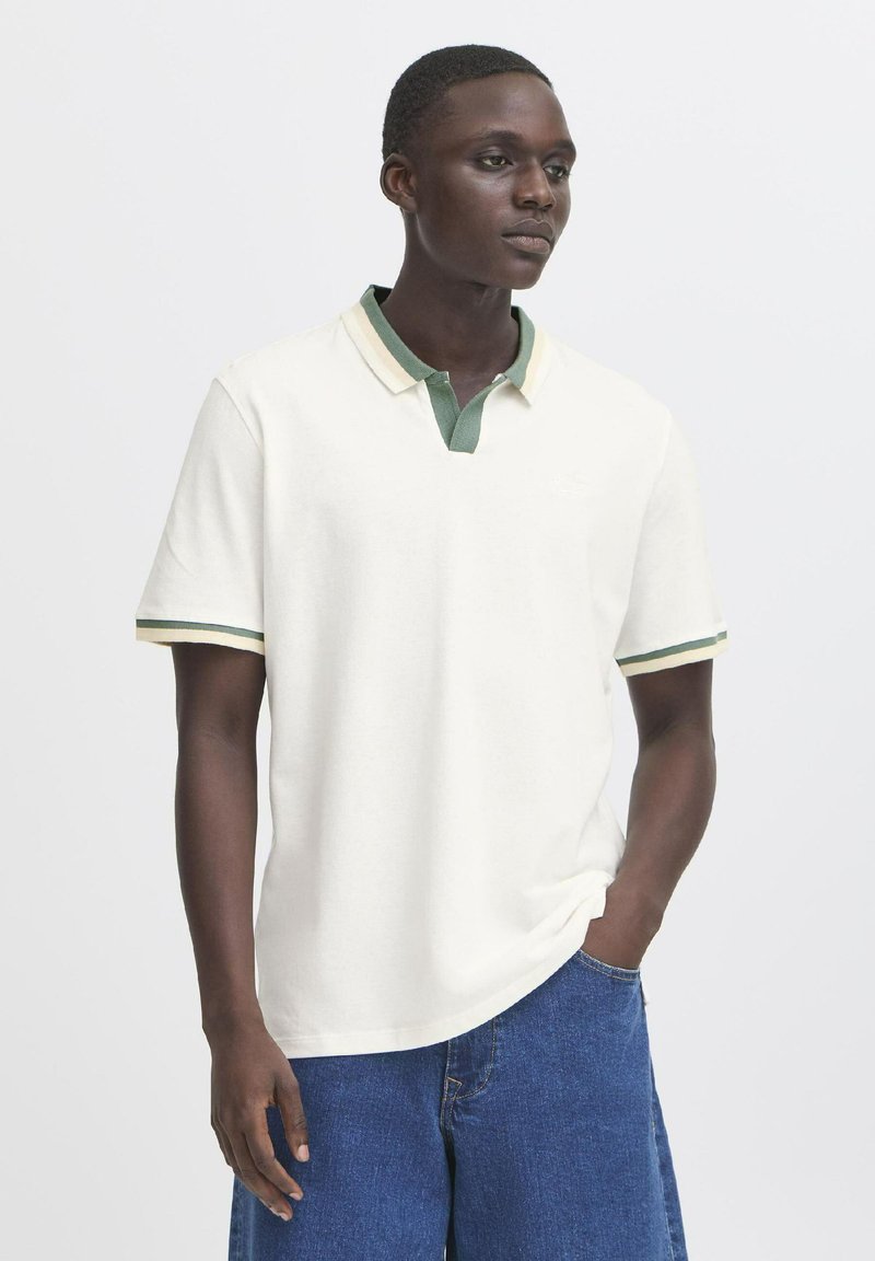 Camisa polo blanca con detalles en verde y amarillo, con cuello abotonado y mangas cortas. Llevada con pantalones cortos de mezclilla azules.