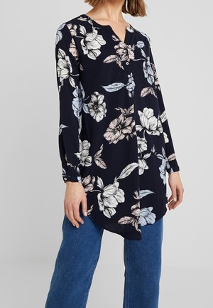 Frau trägt eine marineblaue Langarmbluse mit großen weißen und beigen Blumenmustern und blaue Jeans, steht vor einem schlichten Hintergrund.