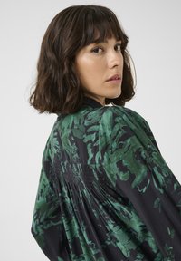 Black garment with green floral pattern, featuring a gathered back, long sleeves, and a relaxed fit. Smooth fabric texture is visible.