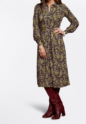 Nainen pukeutunut pitkähihainen, lehtikuvioitu midi-mekko vyöllä, yhdistettynä viininpunaisiin polvipitkkiin nahkasaappaisiin korkokengillä ja kultakoruilla.