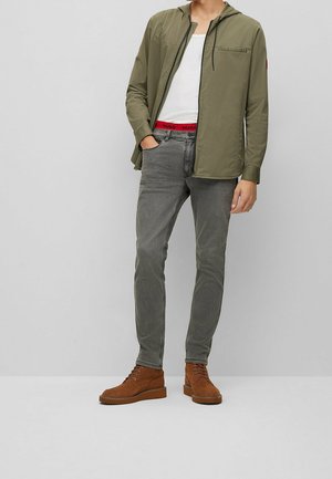 Homme portant une veste à capuche olive, un débardeur blanc, un jean gris et des bottes à lacets en suède marron, se tenant avec une main dans la poche.