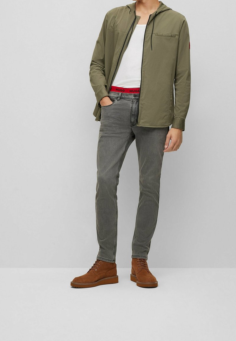 Uomo che indossa una giacca a cappuccio verde oliva, una canotta bianca, jeans grigi e stivaletti in suede marrone con lacci, in piedi con una mano in tasca.