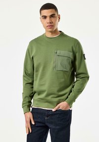 Olivengrüner Sweatshirt mit Rundhalsausschnitt, ausgestattet mit einer Reißverschlusstasche auf der Brust und gerippten Bündchen, kombiniert mit dunklen Jeans.