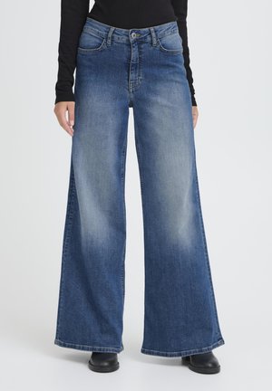 Donna che indossa jeans larghi a vita alta color blu con effetto slavato sul davanti e stivali neri, abbinati a un top nero a maniche lunghe.