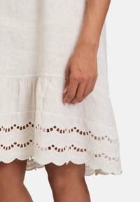 Weißes Leinenkleid mit wellenförmigem Saum, das mit Ausschnitten aus Spitze verziert ist. Der Stoff ist strukturiert und leicht, fällt sanft.