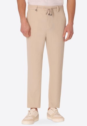 Pantaloni beige a coste con coulisse e chiusura con bottone indossati con sneakers color crema su un modello in piedi su sfondo bianco.