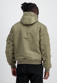 Giacca bomber verde oliva con cappuccio, polsini e fondo a coste. Materiale liscio e lucido con tasche laterali e design dalla vestibilità rilassata.