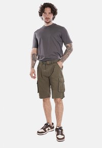 T-shirt grigio a girocollo abbinato a pantaloni cargo verde oliva con multiple tasche e una cintura. Scarpe da ginnastica marroni con dettagli bianchi.